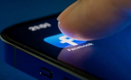 A fingertip pushes on the Facebook App on a mobile phone screen.